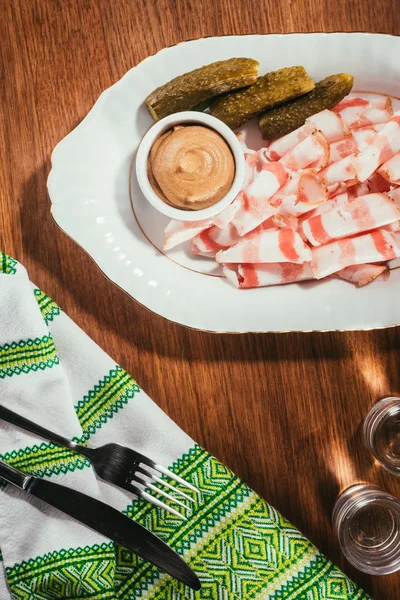 Top view of sliced bacon served with sauce and pickled cucumbers on plate served with drink on wooden table with napkin on wooden table — Stock Photo