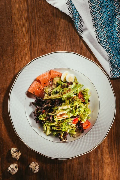 Top view of fresh salad with vegetables, eggs and salmon served on white plate with napkin on wooden table — Stock Photo