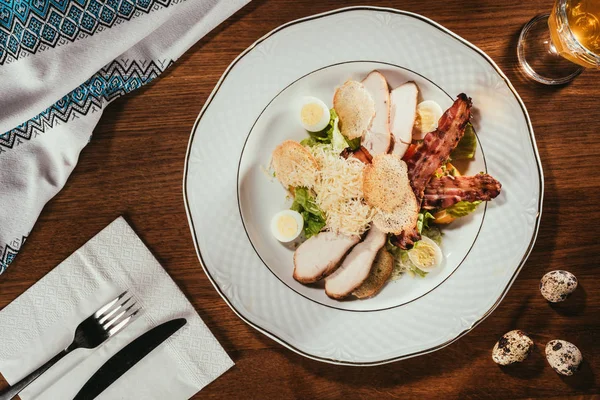 Vista de las lonchas de jamón con carne frita y algunos huevos cocidos en el plato sobre la mesa con tenedor y cuchillo en la servilleta - foto de stock