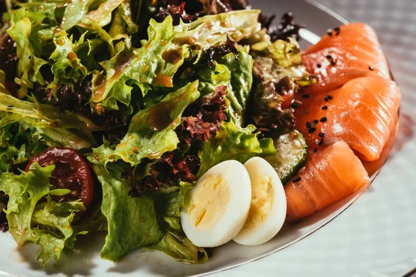 Salad leaves mixed with vegetables laying on plate with some fish slices — Stock Photo