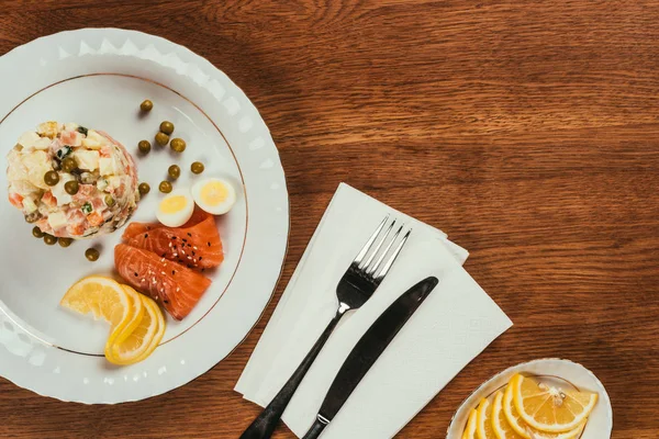 Верхний вид русского салата с лососем и лимоном на белой тарелке на деревянном столе — стоковое фото