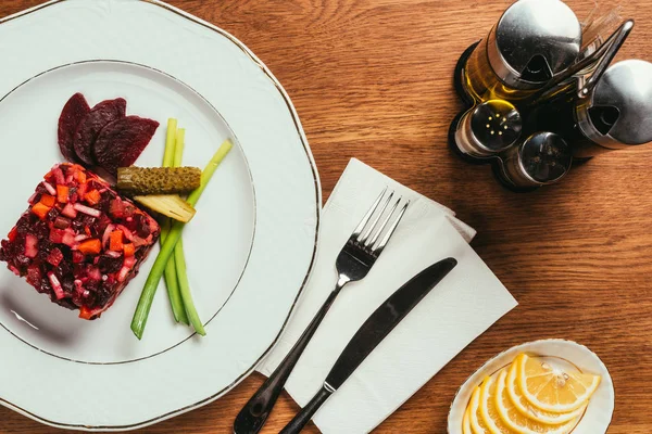 Top view of Vinegret salad served with green onion and pickled cucumber on white plate with condiment dispensers on wooden table — Stock Photo