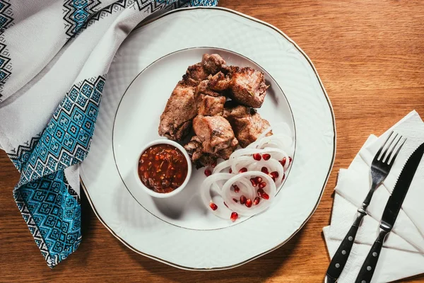 Vista superior da carne grelhada com sementes de cebola e romã na placa branca com guardanapo e talheres na mesa de madeira — Fotografia de Stock