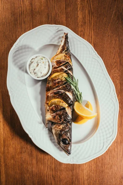Верхний вид печеной рыбы с лимоном и травами на белой тарелке с соусом на деревянном столе — стоковое фото