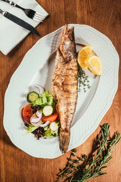 Fried fish laying on white plate with vegetables and orange slices over wooden surface with fork and knife on napkin — Stock Photo