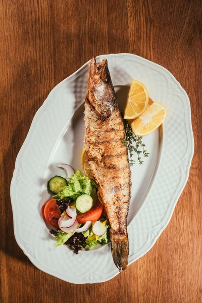 Верхний вид печеной рыбы с лимоном и травами на белой тарелке с салатом на деревянном столе — стоковое фото