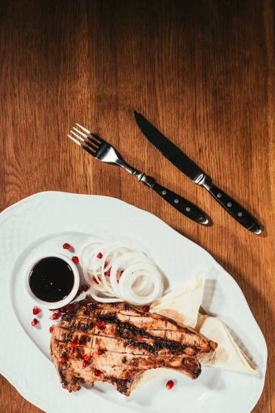 Filete a la parrilla en el plato con aros de cebolla y platillo con salsa sobre la superficie de madera con tenedor y cuchillo - foto de stock