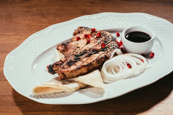 Жареный стейк на тарелке с луковыми кольцами и блюдцем с соусом на деревянной поверхности — стоковое фото
