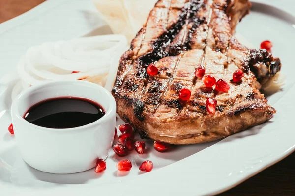 Grilled steak with pomegranate seeds on plate with onion rings and saucer with sauce — Stock Photo