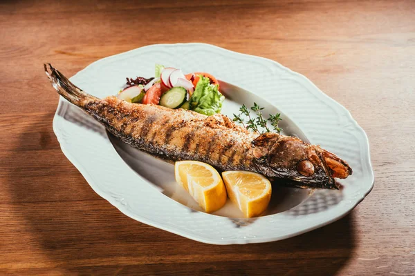 Pesce al forno con limone ed erbe aromatiche su piatto bianco con insalata su tavolo di legno — Foto stock