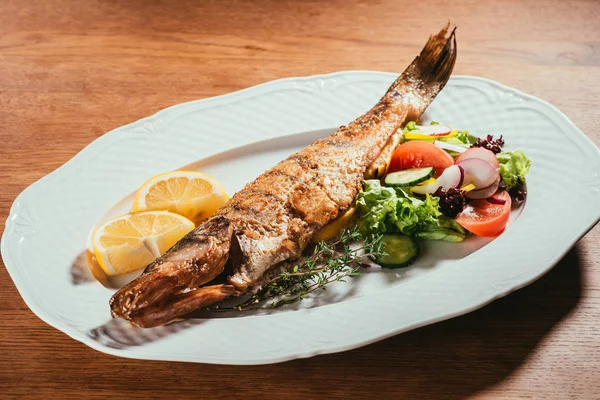 Fried fish laying on white plate with orange slices and sauce over wooden surface — Stock Photo