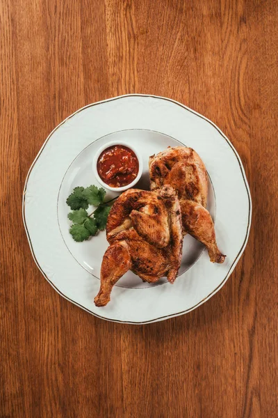 Grilled chicken laying on plate with red sauce in saucer over wooden surface — Stock Photo