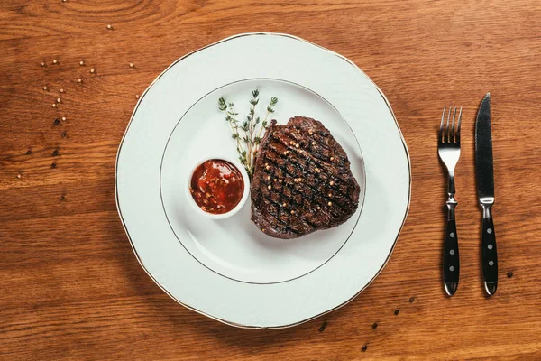 Draufsicht auf gegrilltes Steak mit Pfeffer, Rosmarin und Soße auf weißem Teller auf Holztisch — Stockfoto