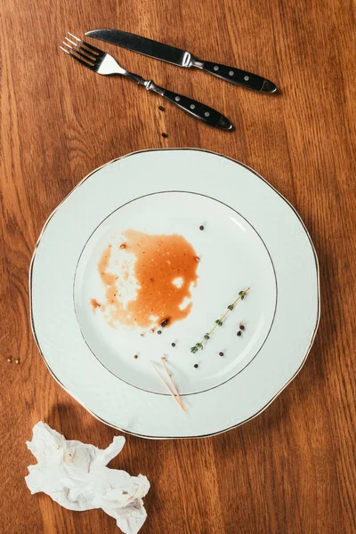 Remainings of meat in almost empty plate on wooden surface with fork, knife and wrinkled napkin — Stock Photo