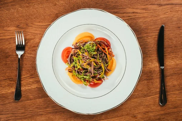 Frisches Gemüse umgeben von Tomaten auf weißem Teller mit Gabel und Messer auf hölzerner Oberfläche geschnitten — Stockfoto