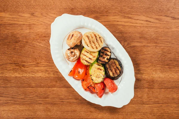 Vista superior de verduras a la parrilla y champiñones en plato blanco sobre mesa de madera - foto de stock