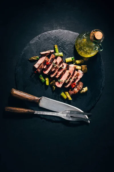 Vista superior de la carne a la parrilla en rodajas gourmet con espárragos, aceite, tenedor de carne y cuchillo en pizarra negra - foto de stock