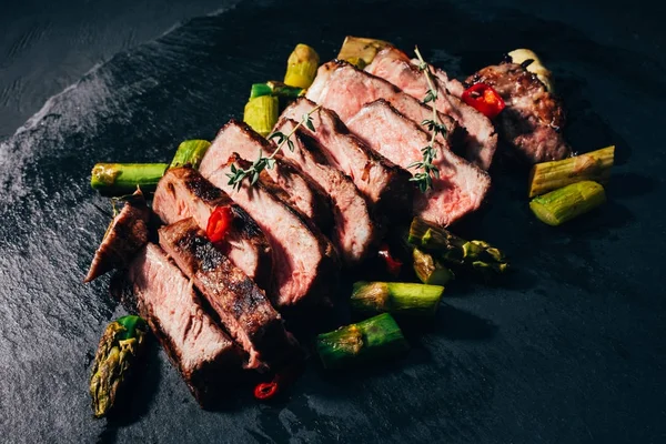 Close-up view of delicious sliced grilled meat with asparagus and spices on black slate board — Stock Photo