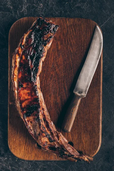 Vue du dessus de délicieuse viande grillée avec couteau sur planche à découper en bois sur noir — Photo de stock