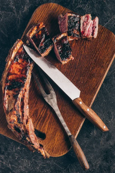 Вид сверху на вкусное мясо на гриле с вилкой и ножом на деревянной доске на черном — стоковое фото