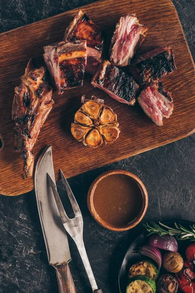 Vista superior de la deliciosa carne a la parrilla con tenedor, cuchillo, salsa y verduras en negro - foto de stock