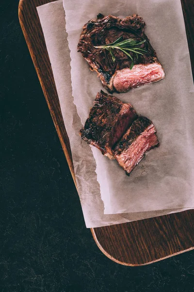 Vue de dessus de la viande grillée délicieuse avec romarin sur papier cuisson et planche de bois sur noir — Photo de stock