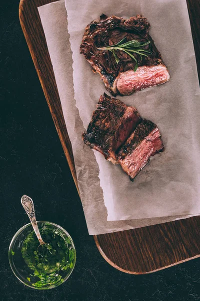 Vue du dessus de délicieuse viande grillée avec romarin sur papier cuisson et sauce dans un bol en verre avec cuillère sur noir — Photo de stock