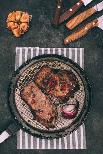 Top view of gourmet roasted steaks on frying pan and various knives on black — Stock Photo