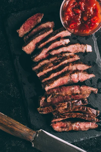 Vista superior de la deliciosa carne a la parrilla en rodajas con salsa y cuchillo en pizarra negra - foto de stock