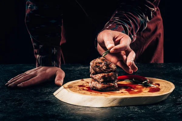 Close-up vue partielle du chef mettant du romarin sur des steaks grillés au piment et sauce sur une planche de bois — Photo de stock