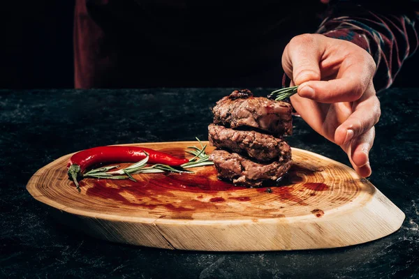 Cropped shot of person putting rosemary on grilled steaks with chili pepper and sauce on wooden board — Stock Photo