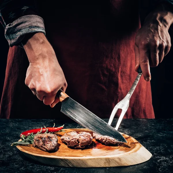 Close-up chef vista parcial em avental com garfo de carne e faca cortando bifes grelhados gourmet com alecrim e pimenta em tábua de madeira — Fotografia de Stock