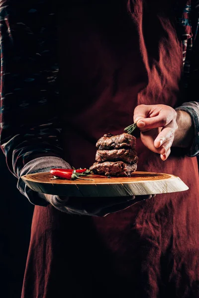 Plan recadré du chef dans un tablier tenant une planche de bois avec de la viande grillée délicieuse, piment et romarin — Stock Photo