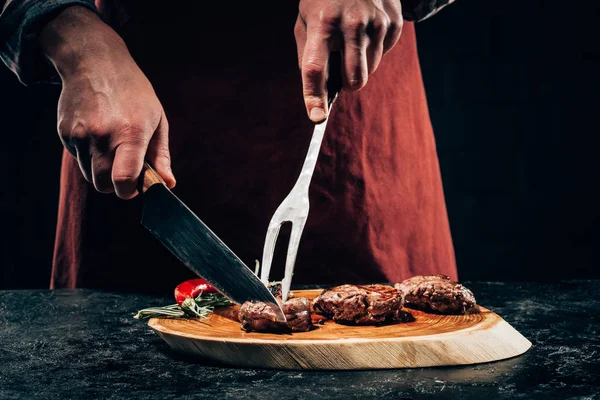 Cropped shot of chef in apron with meat fork and knife slicing gourmet grilled steaks with rosemary and chili pepper on wooden board — Stock Photo