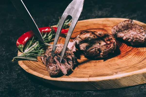 Vista de perto de garfo de carne e faca, deliciosos bifes grelhados com alecrim e pimenta em tábua de madeira — Fotografia de Stock