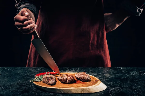 Cropped shot of chef in apron standing with knife and delicious grilled steaks with rosemary and chili pepper on wooden board — Stock Photo