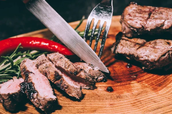 Vista de cerca de la carne a la parrilla en rodajas con romero y chile con tenedor y cuchillo sobre tabla de madera - foto de stock