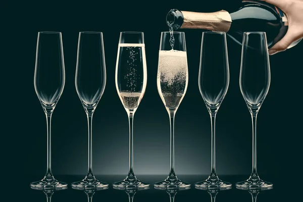 Image recadrée de la femme versant du champagne de la bouteille dans six verres transparents sur noir — Photo de stock