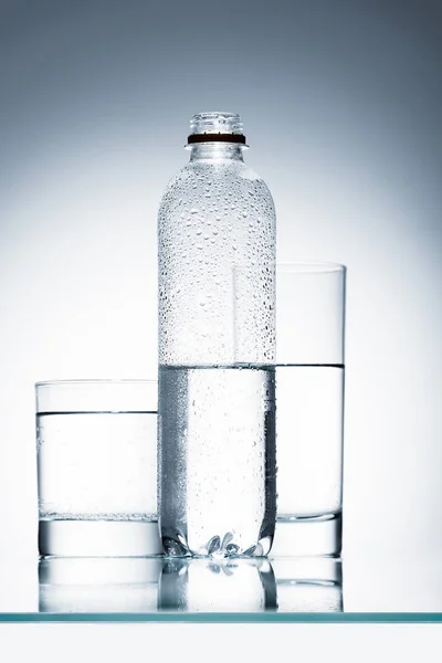 Botella de plástico y vasos de agua limpia en la superficie reflectante - foto de stock