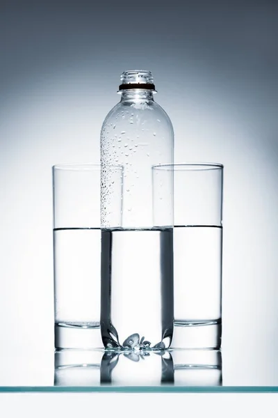 Пластиковая бутылка и стаканы чистой воды на отражающей поверхности — стоковое фото