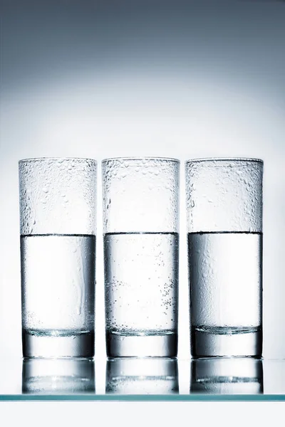 Vasos medio llenos de agua en fila en la superficie reflectante - foto de stock