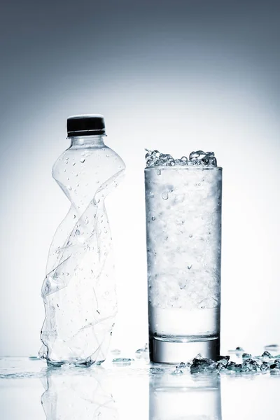 Vidro de água fria com gelo e garrafa de plástico amassado na superfície reflexiva em branco — Fotografia de Stock