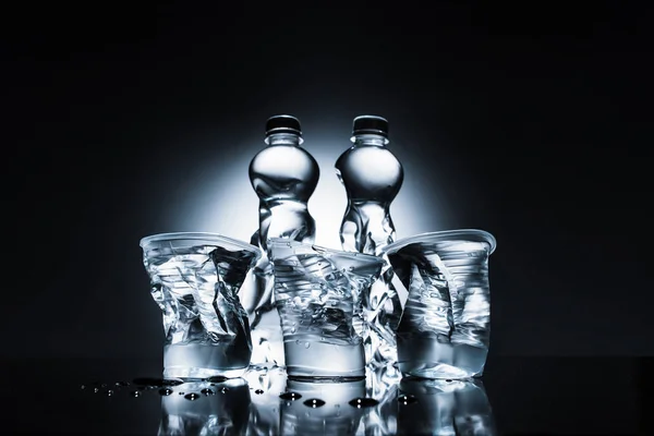 Botellas de plástico arrugado y tazas con agua en la oscuridad - foto de stock