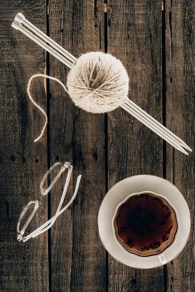Vista superior de agujas de punto, hilo, taza de té y anteojos sobre fondo de madera - foto de stock