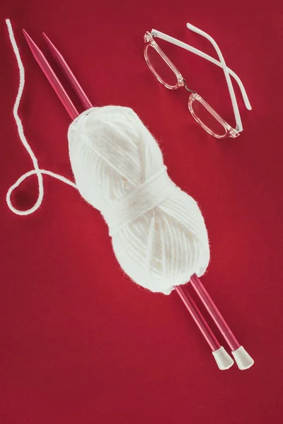 Vue de dessus des lunettes, boule de fil blanc et aiguilles à tricoter, isolé sur rouge — Photo de stock