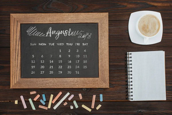 Vista superior da xícara arranjada de café, caderno vazio, giz colorido e quadro com calendário de agosto — Fotografia de Stock