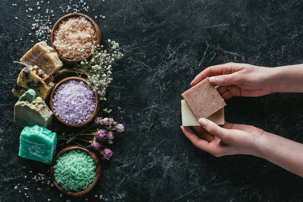 Vue recadrée des mains féminines avec savon naturel fait maison, fleurs séchées et sel de mer sur la surface de marbre noir — Photo de stock