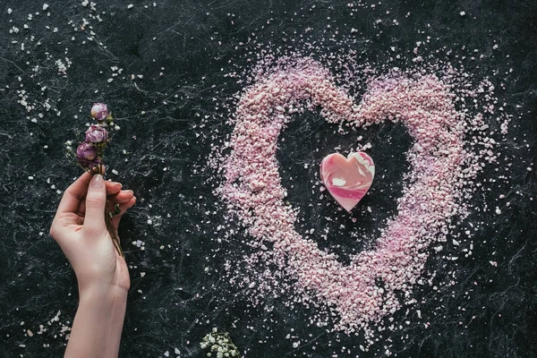 Geschnittene Ansicht der weiblichen Hand mit getrockneten Rosen auf schwarzer Marmoroberfläche mit rosa Herzseife und Meersalz für den Wellnessbereich am Valentinstag — Stockfoto