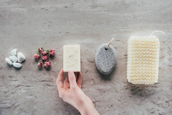 Vista recortada de la mano con piedras, rosas secas, jabón natural, piedra pómez y esponja de baño para spa en superficie de mármol - foto de stock