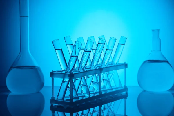 Tubi di vetro con liquido sul supporto e fiaschette di vetro sul tavolo su blu — Foto stock
